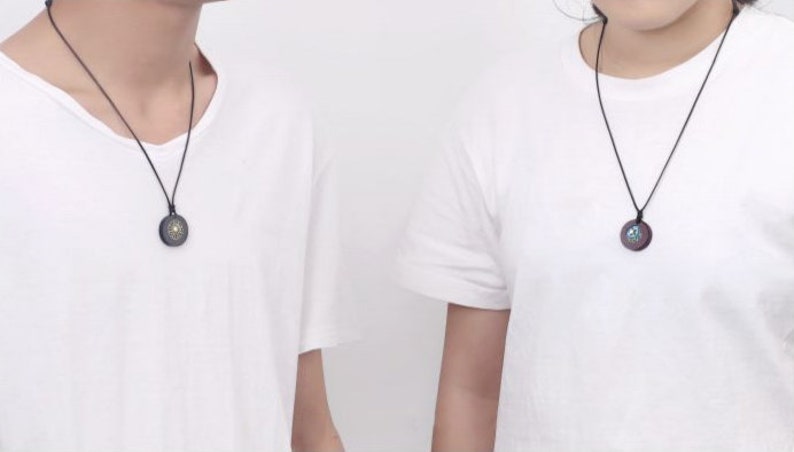 May include: Two people wearing white t-shirts with black necklaces and round pendants. The pendants have a dark wood finish with a gold design.
