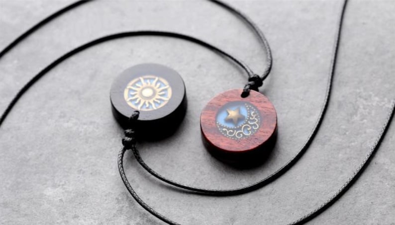 May include: Two wooden necklaces with pendants. One pendant is black with a gold sun design, the other is red with a blue moon and gold star design.
