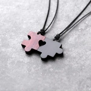 May include: Two black cord necklaces with a wooden puzzle piece pendant. The puzzle pieces fit together to form a heart shape.