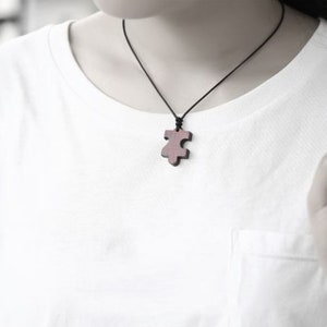 May include: A wooden puzzle piece pendant necklace on a black cord. The pendant is a dark brown color and has a slightly reddish hue.