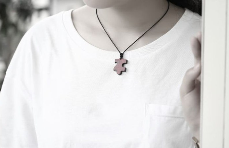 May include: A wooden puzzle piece pendant necklace on a black cord. The pendant is a dark brown color and has a slightly reddish hue.