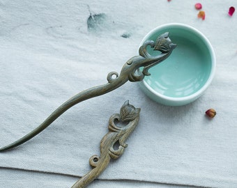 May include: Two wooden hair sticks with intricate carvings of stylized cats. The sticks are dark brown and have a smooth, polished finish. The sticks are lying on a light gray fabric background.