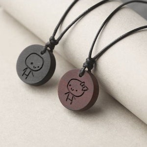 May include: Two wooden necklaces with black cords. Each pendant is a round disc with a simple stick figure design carved into the wood. One pendant is black and the other is brown.