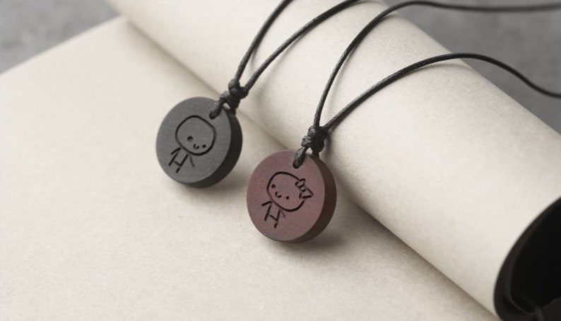 May include: Two wooden necklaces with black cords. Each pendant is a round disc with a simple stick figure design carved into the wood. One pendant is black and the other is brown.