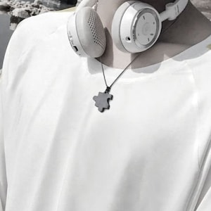 May include: A white long-sleeved shirt with yellow stripes on the sleeves. The shirt has a round neckline and a black puzzle piece pendant necklace. The person is wearing white headphones.