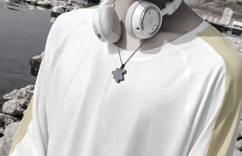 May include: A white long-sleeved shirt with yellow stripes on the sleeves. The shirt has a round neckline and a black puzzle piece pendant necklace. The person is wearing white headphones.
