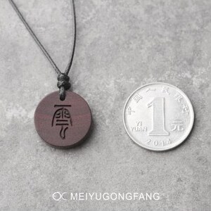May include: A dark brown wooden pendant necklace with a black cord. The pendant has a Chinese character engraved on it. The necklace is displayed on a gray surface next to a Chinese yuan coin.