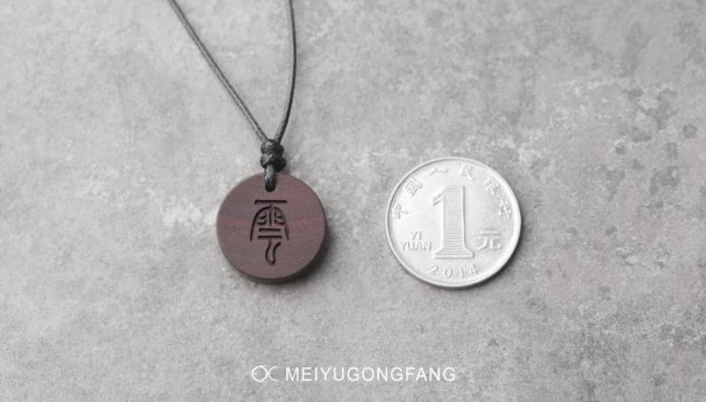 May include: A dark brown wooden pendant necklace with a black cord. The pendant has a Chinese character engraved on it. The necklace is displayed on a gray surface next to a Chinese yuan coin.