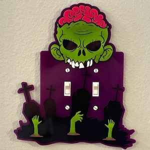 May include: A decorative light switch cover featuring a zombie design. The zombie has a green face, pink brain, and white teeth. The purple cover has three light switches, tombstones, and green zombie hands.