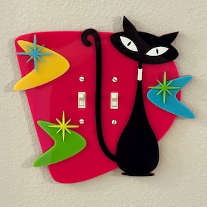 Könnte beinhalten: Eine Lichtschalterabdeckung im Retro-Stil mit einer schwarzen Katzen-Silhouette mit weißen Augen, vor einem rosa Hintergrund. Sie zeigt Stern- und Bumerangformen in Gelb, Grün und Blau. Das Design hat einen Mid-Century-Modern-Stil.