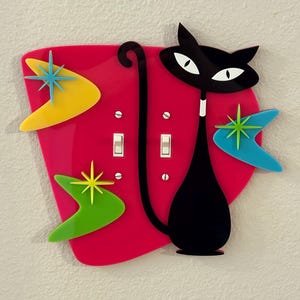 Op de afbeelding: Een retro lichtschakelaar cover met een zwart katten silhouet met witte ogen, tegen een felroze achtergrond. Het heeft ster- en boemerang accenten in geel, groen en blauw, die een mid-century modern esthetiek oproepen.