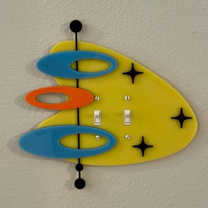 May include: A retro-style light switch cover with a yellow, boomerang-shaped base. It features blue and orange oval accents, black starbursts, and two toggle switches. The design includes a vertical black line with black circles.