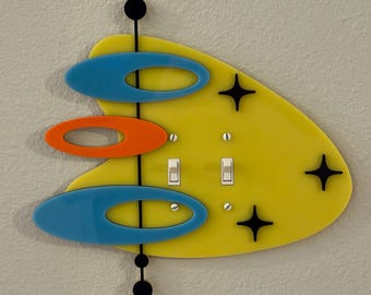 May include: A retro-style light switch cover with a yellow, boomerang-shaped base. It features blue and orange oval accents, black starbursts, and two toggle switches. The design includes a vertical black line with black circles.