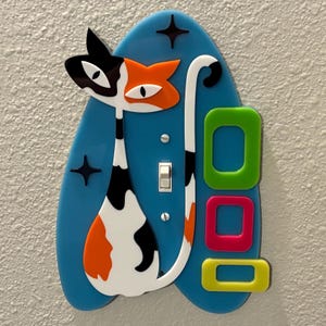 May include: A whimsical light switch cover featuring a stylized calico cat design. The cat is black, white, and orange, set against a blue background with starburst accents. Rectangular shapes in green, pink, and yellow add a pop of color.