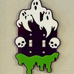 May include: A purple and green Halloween-themed light switch cover. The cover features white ghost and skull cutouts, with green slime dripping from the bottom. The cover has two light switch openings.