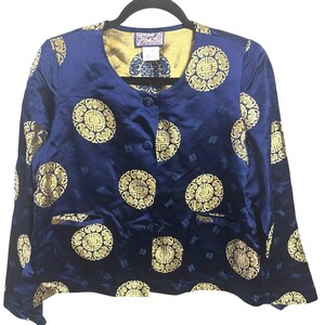 May include: A navy blue jacket with a gold circular pattern design. The jacket has a round neckline, three buttons, and two front pockets. The interior lining is gold. The jacket is hanging on a black hanger.