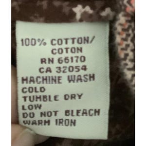 Puede incluir: Primer plano de una etiqueta blanca con texto negro. La etiqueta dice: "100% COTTON/COTON, RN 66170, CA 32054, LAVAR A M&Aacute;QUINA EN FR&Iacute;O, SECADORA A BAJA TEMPERATURA, NO USAR LEJ&Iacute;A, PLANCHAR A BAJA TEMPERATURA." La etiqueta est&aacute; sobre una tela marr&oacute;n.