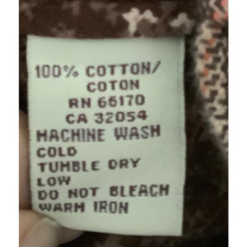 Puede incluir: Primer plano de una etiqueta blanca con texto negro. La etiqueta dice: "100% COTTON/COTON, RN 66170, CA 32054, LAVAR A M&Aacute;QUINA EN FR&Iacute;O, SECADORA A BAJA TEMPERATURA, NO USAR LEJ&Iacute;A, PLANCHAR A BAJA TEMPERATURA." La etiqueta est&aacute; sobre una tela marr&oacute;n.