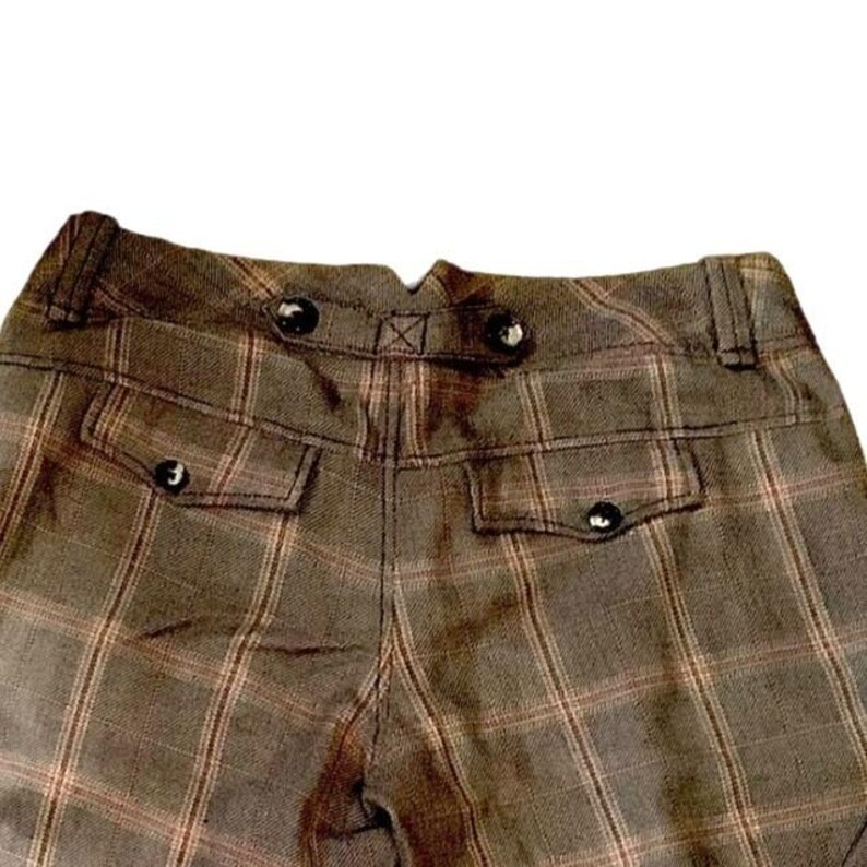 Puede incluir: Primer plano de la parte trasera de unos pantalones a cuadros marrones. Los pantalones tienen dos bolsillos con cierre de bot&oacute;n y botones decorativos en la cintura. El estampado de cuadros es marr&oacute;n y beige.