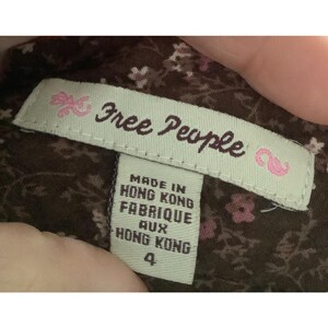 Puede incluir: Primer plano de una etiqueta de ropa de la marca "Free People". La etiqueta es rectangular con el nombre de la marca en cursiva. Debajo, una etiqueta m&aacute;s peque&ntilde;a con el texto "Made in Hong Kong, Fabrique aux Hong Kong, 4". El fondo es un estampado floral.
