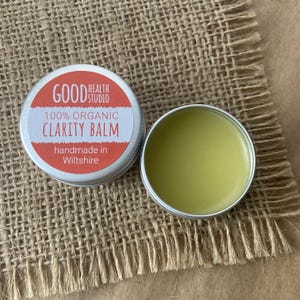 May include: A small silver tin with a red and white label that reads "GOOD HEALTH STUDIO 100% ORGANIC CLARITY BALM handmade in Wiltshire". The tin is open and contains a yellow balm.