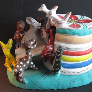 Könnte beinhalten: Eine farbenfrohe Keramikfigur, die eine Gruppe von Tieren und eine Person zeigt, die sich um eine mehrschichtige Struktur versammelt haben. Die Struktur ist mit Streifen in Rot, Gelb, Blau und Braun bemalt. Die Tiere sind in verschiedenen Farben bemalt, darunter Weiß, Schwarz, Braun und Gelb. Die Figur ist eine skurrile und verspielte Darstellung einer Gemeinschaft.