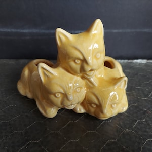 Puede incluir: Un conjunto de tres figuras de maceta de gato de cerámica amarilla. Los gatos están apilados uno encima del otro, con el gato más grande en la parte inferior y el gato más pequeño en la parte superior. Los gatos tienen rasgos realistas, incluidos ojos, narices y bocas.