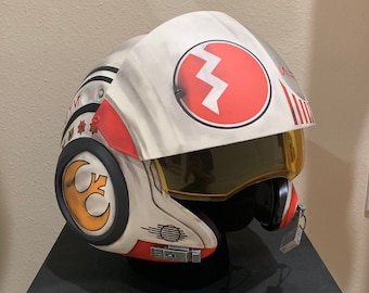 May include: A white and red pilot helmet with a yellow visor and a red circle with a lightning bolt on the top. The helmet has a Rebel Alliance symbol on the side.