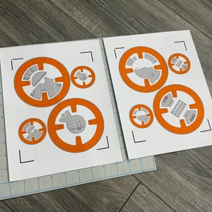 Könnte beinhalten: Zwei Papierbögen mit orangefarbenen und grauen BB-8-Droiden-Ausschnitten. Die Designs zeigen kreisförmige Formen mit detaillierten Abschnitten, ideal für Bastel- oder Themenprojekte. Die Bögen liegen auf einer grauen Schneidematte.