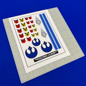 May include: A sheet of white paper with blue, red, yellow, and black decals for a model spaceship. The decals include the Rebel Alliance symbol, a starfighter symbol, and a series of stripes. The text on the sheet reads "Yavin Pilot Decals by Moonsault".