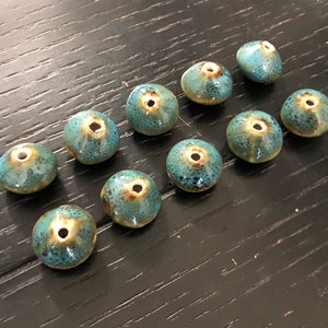 Könnte beinhalten: Zwölf tealblaue Keramikperlen mit gesprenkelter Oberfläche und einem kleinen Loch in der Mitte. Die Perlen sind in zwei Reihen auf einer dunklen Holzoberfläche angeordnet.