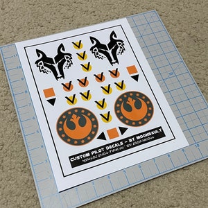 May include: A sheet of custom pilot decals featuring a black wolf head, a Rebel Alliance symbol, and various other symbols in orange, yellow, and black. The text "CUSTOM PILOT DECALS - BY MOONSAULT" is printed at the bottom.