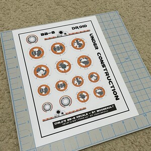 May include: A sheet of white paper with a set of 12 orange and gray BB-8 droid decals. The decals are labeled "Child's BB-8 Decals - By Moonsault".