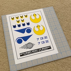 May include: A sheet of white paper with black and white decals for a Star Wars Rey helmet. The decals include the Rebel Alliance symbol, a blue and white symbol, and the numbers 733. The text "REY'S HELMET DECALS - BY MOONSAULT" is printed at the bottom of the sheet.