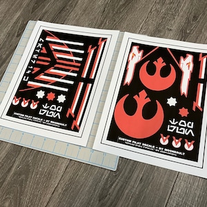 Op de afbeelding: Twee vellen met zwarte en witte aangepaste piloot-stickers met rode accenten. De stickers bevatten het Rebel Alliance-symbool, de tekst "CUSTOM PILOT DECALS - BY MOONBAULT" en andere geometrische vormen.