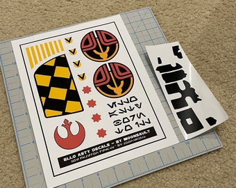 May include: A set of Star Wars themed vinyl decals featuring a Rebel Alliance symbol, a checkered pattern, and two circular symbols with red, yellow, and black accents. The text "ELLO ASTY DECALS - BY MOONSAULT GOVM 7X2ИТОЙ РИЧКIN SV COOK" is printed on the sheet.