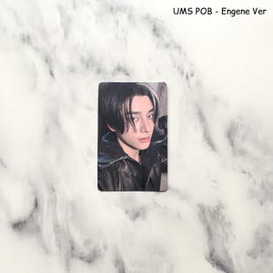 Op de afbeelding: Een rechthoekige fotokaart met een persoon met zwart haar en een witte lok, gekleed in een zwarte leren jas. De kaart heeft bovenaan de tekst "UMS POB - Engene Ver".