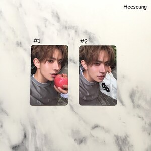 May include: Two rectangular cards featuring a person. Card #1 shows the person holding a red apple. Card #2 shows the person holding a white garment with a black design. The name "Heeseung" is printed at the top right.