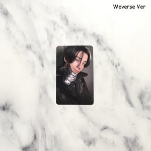 Op de afbeelding: Een rechthoekige fotokaart met een persoon met donker haar en een zwarte jas. De persoon glimlacht en houdt een hand omhoog met een gedetailleerde, metalen handschoen. De tekst "Weverse Ver" staat rechtsboven.