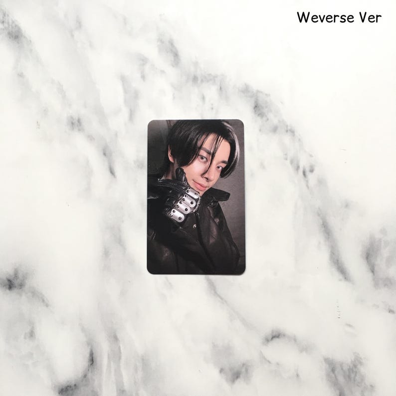 Op de afbeelding: Een rechthoekige fotokaart met een persoon met donker haar en een zwarte jas. De persoon glimlacht en houdt een hand omhoog met een gedetailleerde, metalen handschoen. De tekst "Weverse Ver" staat rechtsboven.