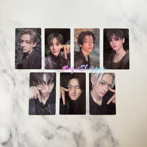 May include: Seven rectangular trading cards featuring individual portraits of young people with dark hair, some wearing black jackets. The cards are arranged in two rows, with the text MagicShoppen written in pink cursive script across the center.