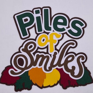 May include: A brown and green paper craft with the words "Piles of Smiles" in a white outline. The craft features a fall theme with brown, yellow, and red leaves.