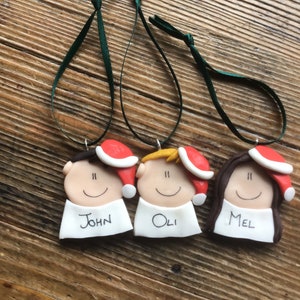 May include: Three personalized Christmas ornaments with red Santa hats, white faces, and black hair. The ornaments are labeled "John", "Oli", and "Mel".