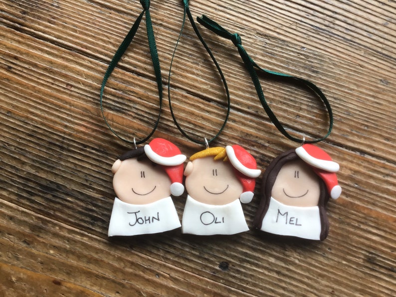 May include: Three personalized Christmas ornaments with red Santa hats, white faces, and black hair. The ornaments are labeled "John", "Oli", and "Mel".