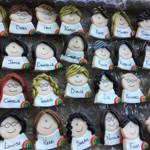 Op de afbeelding: Een verzameling van 24 gepersonaliseerde taarttoppers met kleurrijke regenboogaccenten. Elke topper heeft een cartoongezicht met een naam eronder geschreven. De namen zijn Christine, Debs, Ian, Karen, Anne, Morag, Shona, Katie, Jamie, Danielle, Kimberley, Fiona, Kirstin, Eilidh, Anna, Charlie, Ingrid, Diane, Liz, Richard, Morven, Louise, Kerri, Sarah, Nicola, Fiona en Chris.