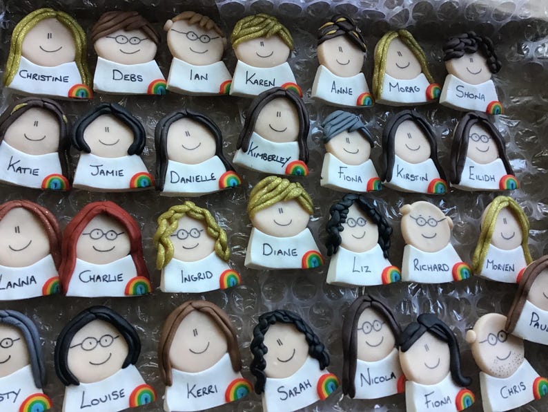 Op de afbeelding: Een verzameling van 24 gepersonaliseerde taarttoppers met kleurrijke regenboogaccenten. Elke topper heeft een cartoongezicht met een naam eronder geschreven. De namen zijn Christine, Debs, Ian, Karen, Anne, Morag, Shona, Katie, Jamie, Danielle, Kimberley, Fiona, Kirstin, Eilidh, Anna, Charlie, Ingrid, Diane, Liz, Richard, Morven, Louise, Kerri, Sarah, Nicola, Fiona en Chris.