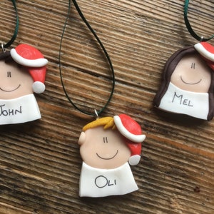 May include: Three personalized Christmas ornaments with red Santa hats, each with a different name: John, Mel, and Oli. The ornaments are made of white clay with black eyes and a smiling mouth.