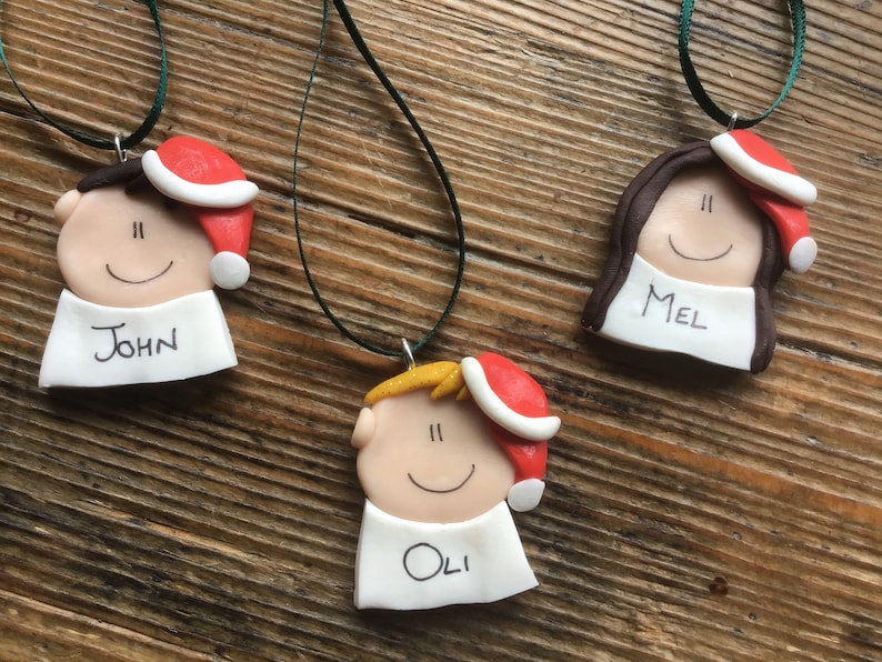 May include: Three personalized Christmas ornaments with red Santa hats, each with a different name: John, Mel, and Oli. The ornaments are made of white clay with black eyes and a smiling mouth.