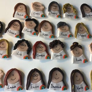 Op de afbeelding: Een verzameling van 24 gepersonaliseerde naamstickers met cartoongezichten met verschillende kapsels en brillen. Elke sticker heeft een regenboogontwerp en een naam in het zwart.