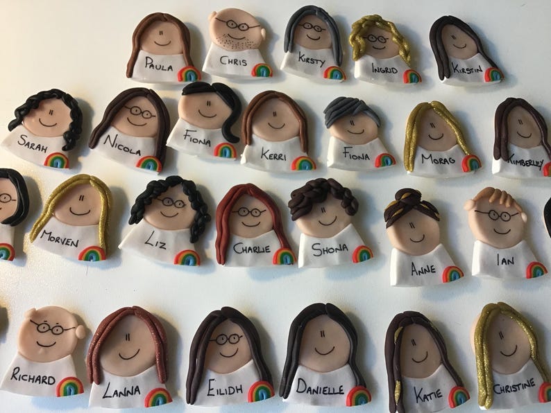 Op de afbeelding: Een verzameling van 24 gepersonaliseerde naamstickers met cartoongezichten met verschillende kapsels en brillen. Elke sticker heeft een regenboogontwerp en een naam in het zwart.
